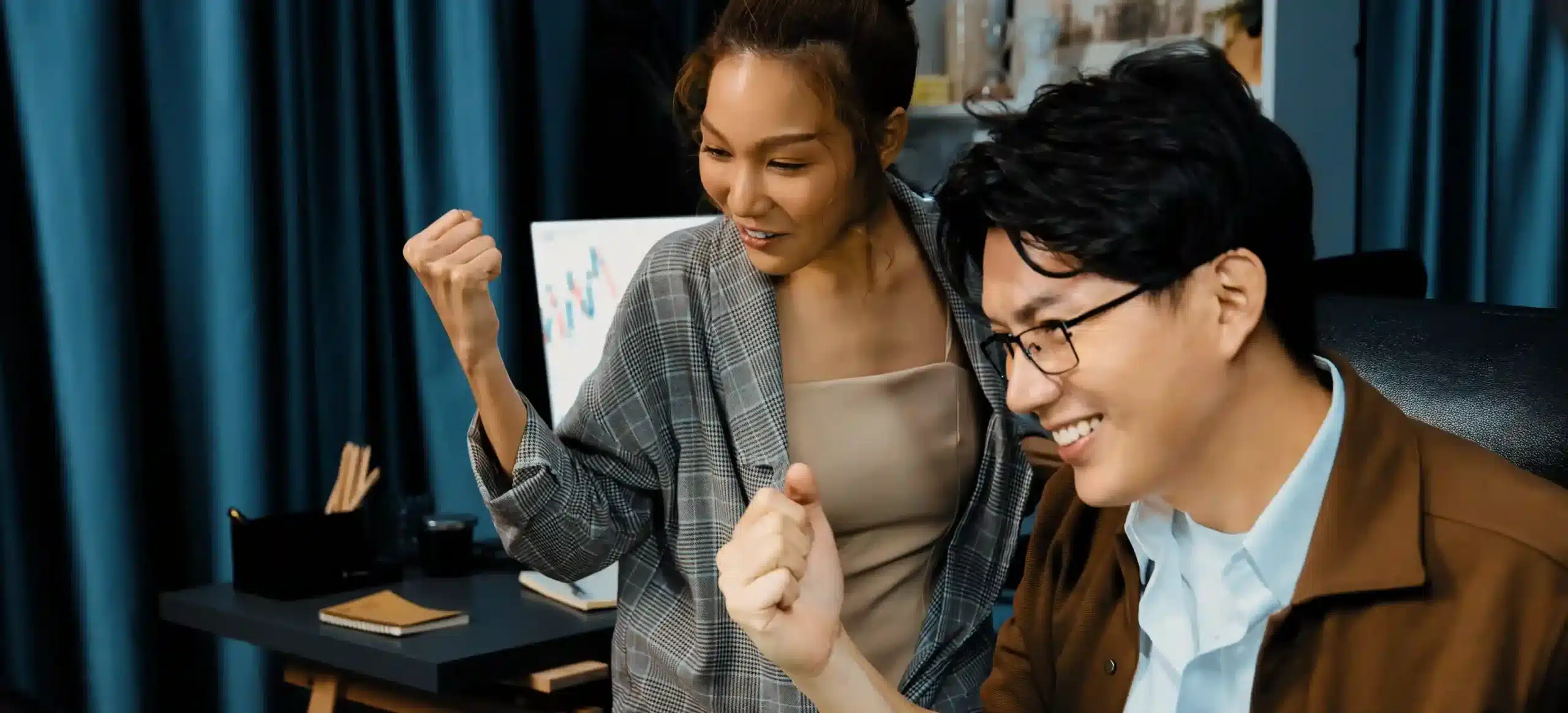 Two colleagues in an office celebrating a success while looking at a computer screen. Both are smiling and raising their fists excitedly, with charts and office supplies visible in the background