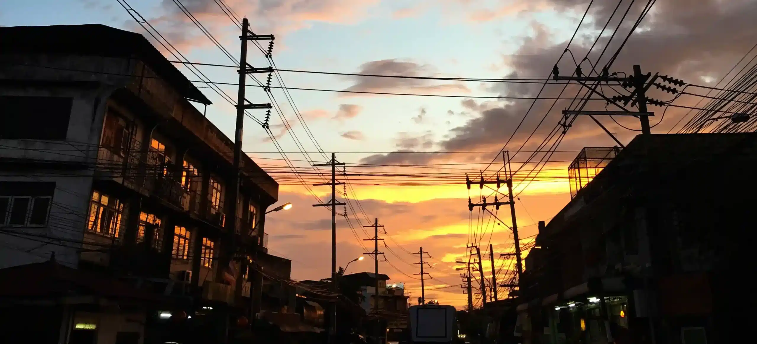 A city street at sunset with silhouettes of buildings and numerous overhead power lines against an orange and pink sky.