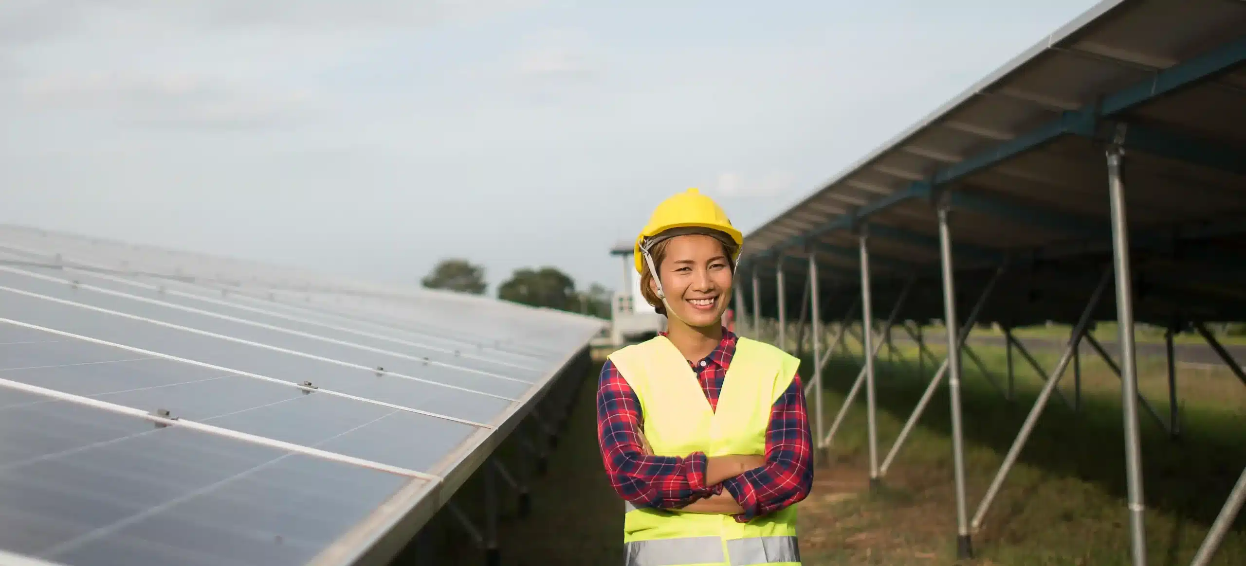 A smiling solar power engineer wearing a yellow hard hat and reflective safety vest stands with folded arms between rows of large outdoor solar panels on a sunny day.