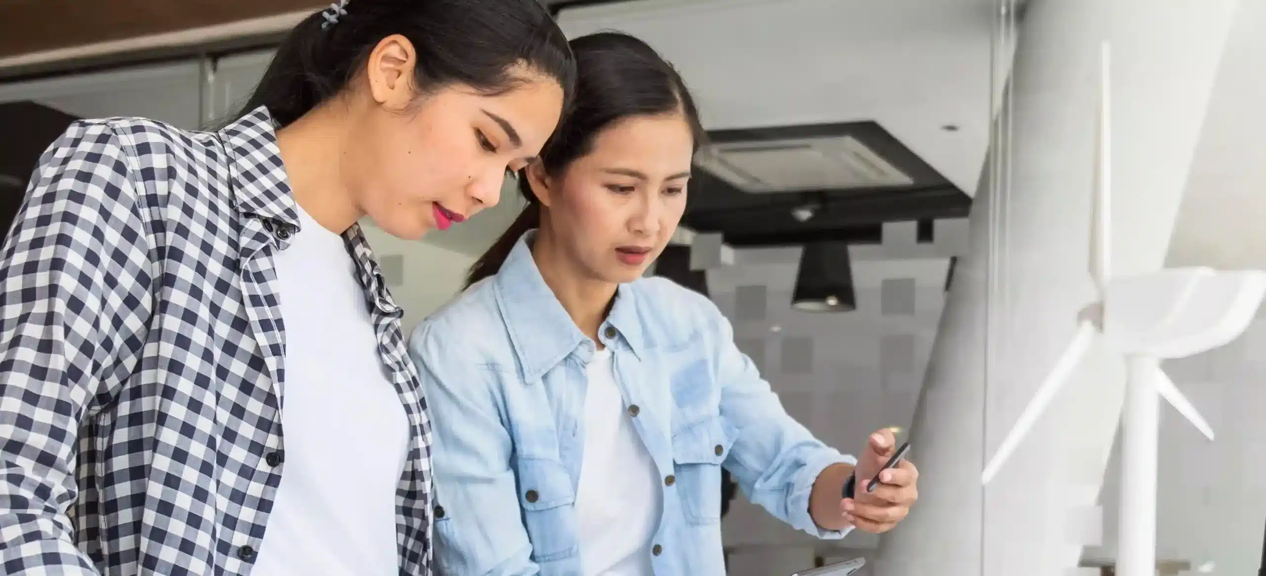 Two women working together indoors, looking down at documents or a device. One wears a checkered shirt and the other a light denim shirt. A small wind turbine model is visible in the background.